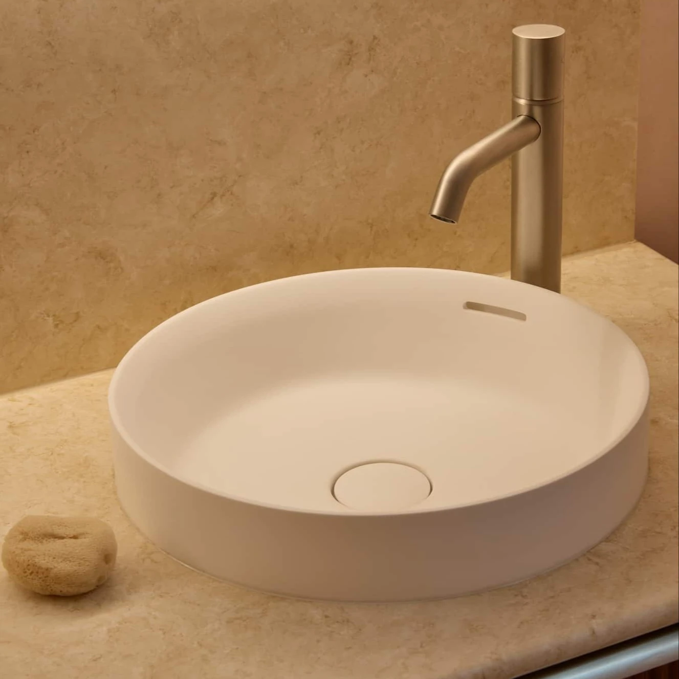 Modern bathroom sink with white circular basin and brushed gold faucet on beige marble countertop.