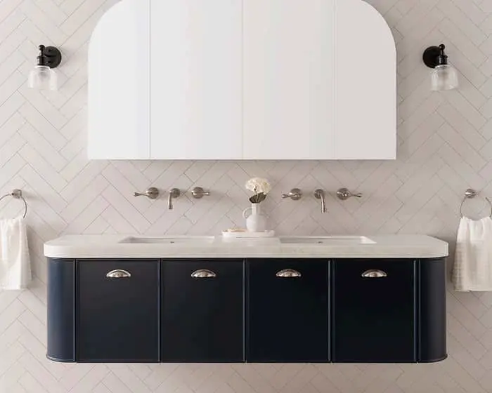 Modern bathroom with navy blue vanity, white double sinks, arched mirror cabinet, and herringbone tile wall.
