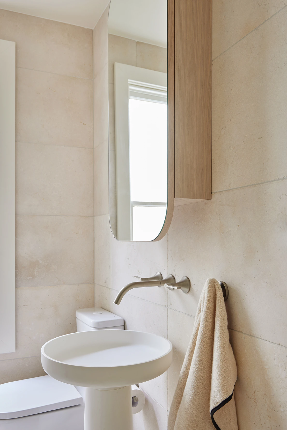 Small bathroom with beige stone tiles, a white pedestal sink, wall-mounted brushed nickel faucet, tall mirror, and a beige towel on a hook.