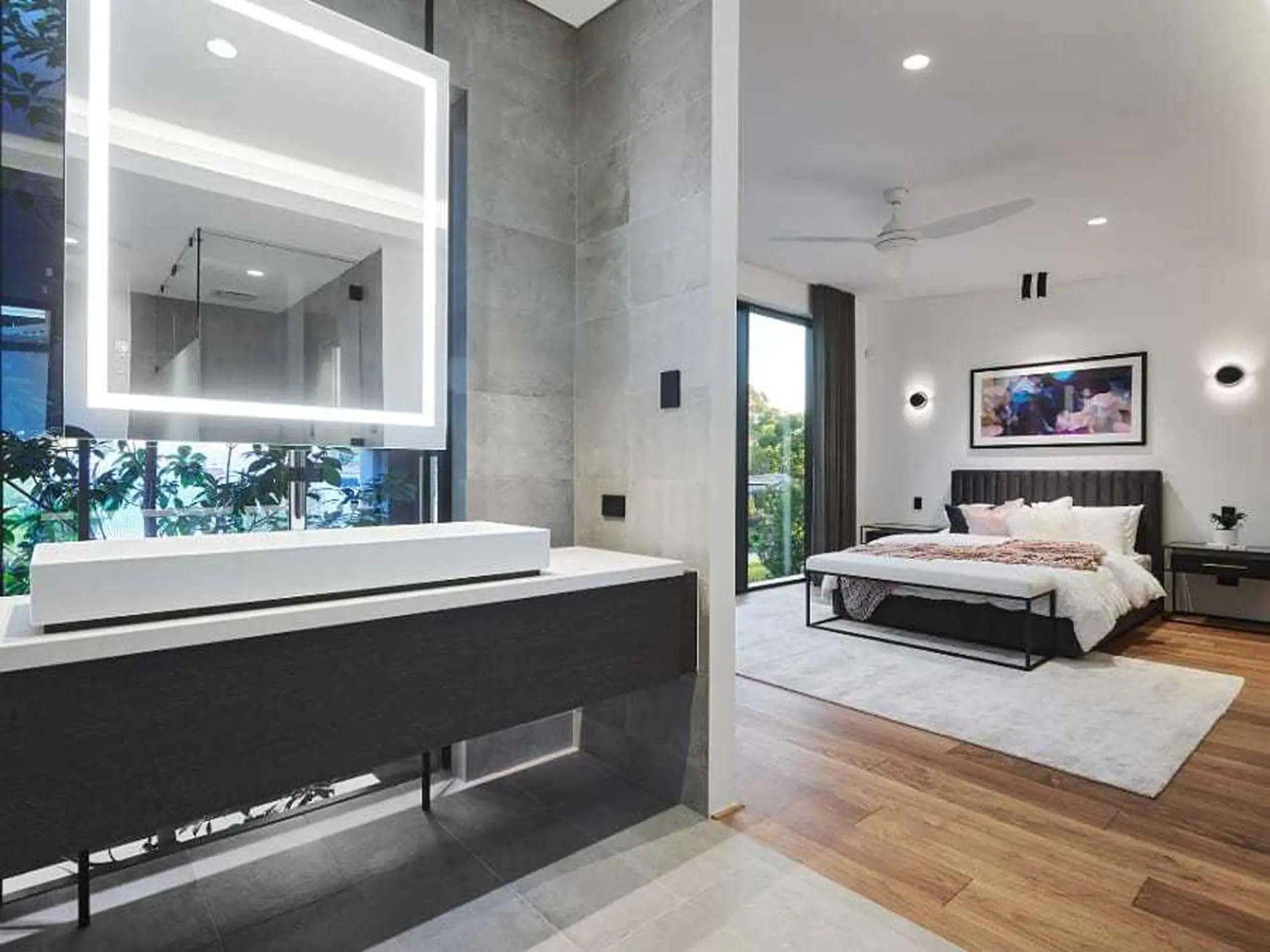 Modern bedroom with ensuite bathroom featuring illuminated mirror, dark vanity, gray tile, and king bed with white linens.