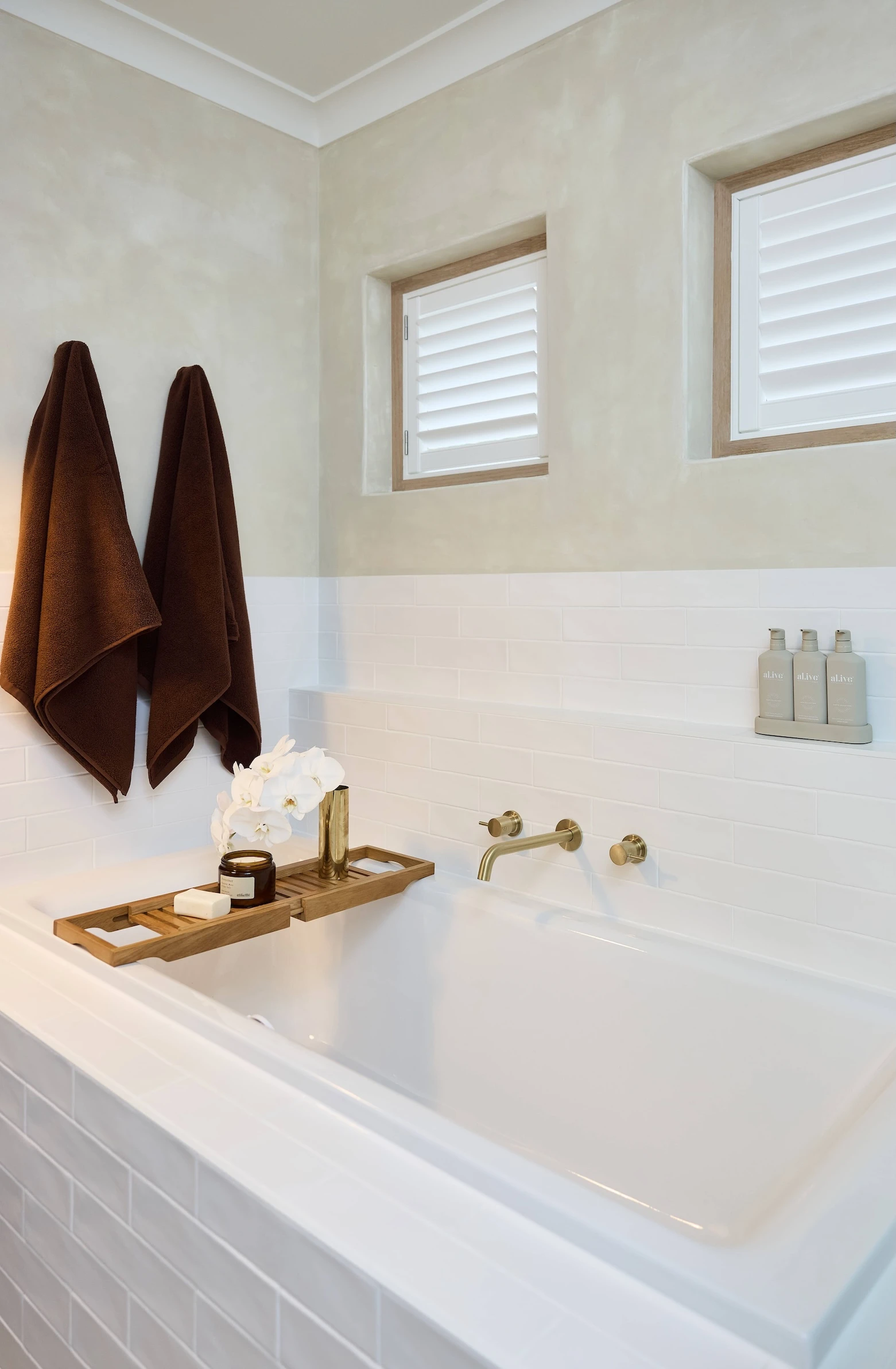 Modern bathroom with white bathtub, wooden tray holding orchids, brown towels, and brass fixtures under shuttered windows.
