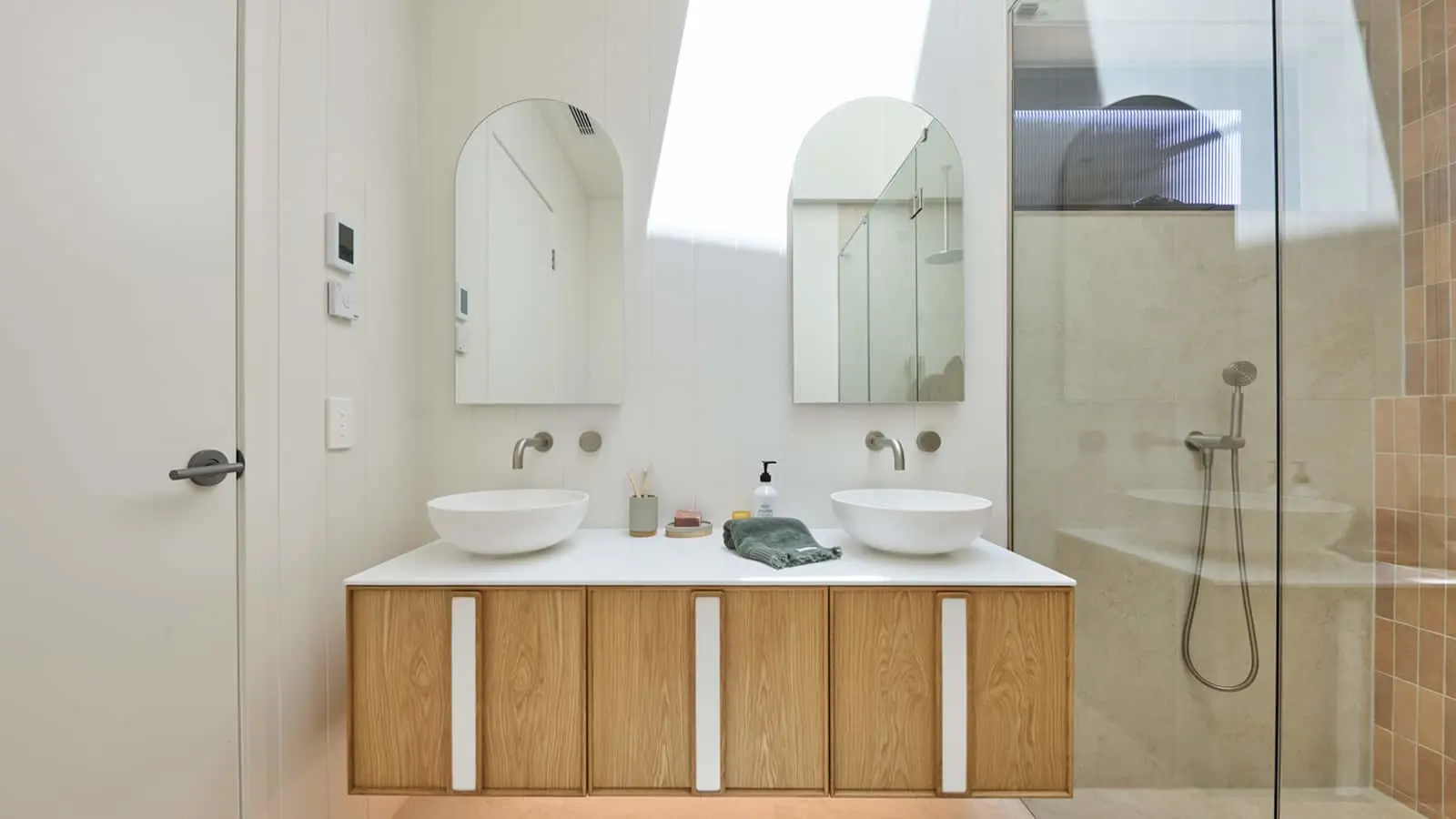 Modern bathroom with dual white vessel sinks on wooden vanity, arched mirrors, and glass shower enclosure.