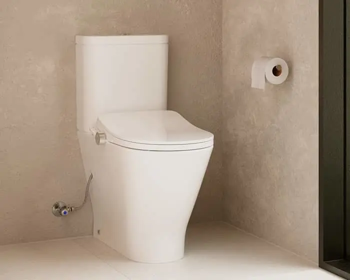 Modern white toilet with closed lid in a minimalist bathroom with beige textured walls and toilet paper mounted nearby.