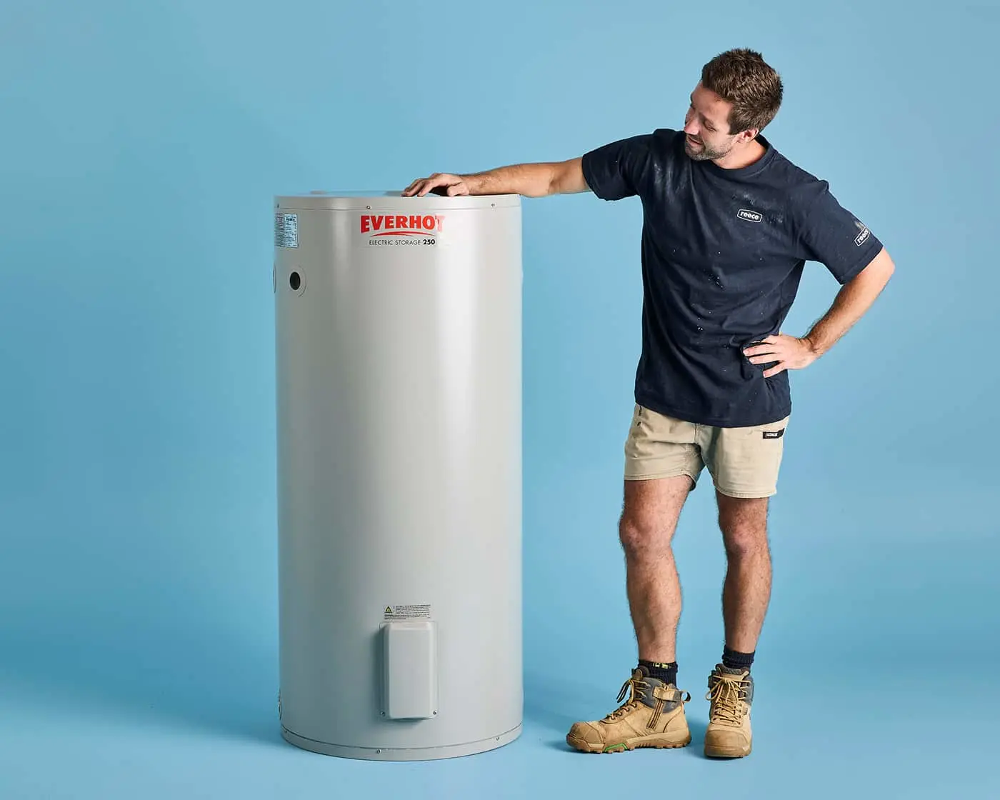 Person in navy shirt and work boots examining an Everhot electric storage water heater against a blue background.