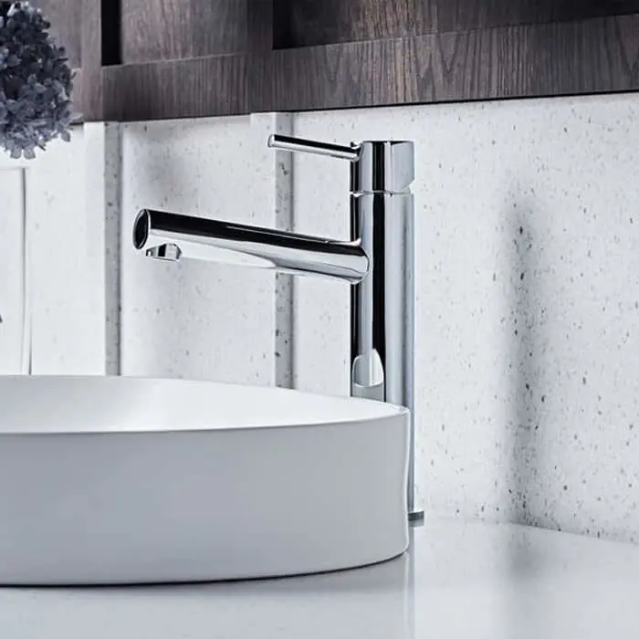 Modern chrome bathroom faucet with white vessel sink against textured white wall and dark wood cabinetry.