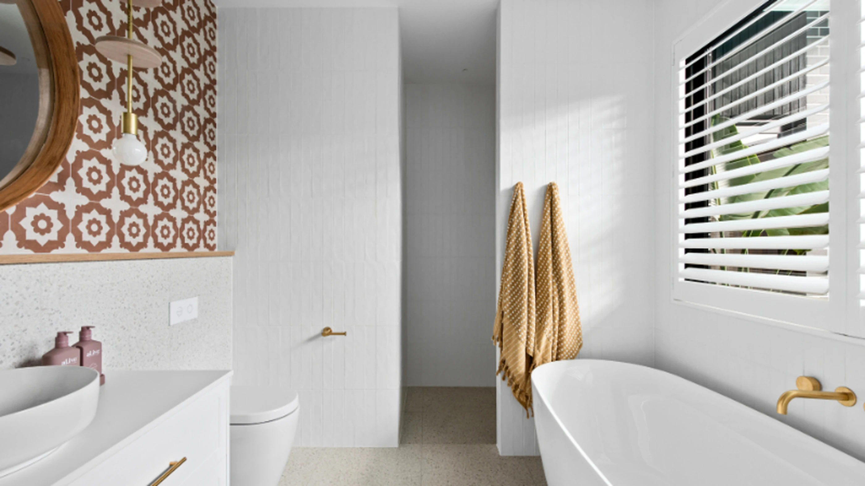 Modern white bathroom with patterned accent wall, gold fixtures, white bathtub, and yellow towels hanging near window blinds.