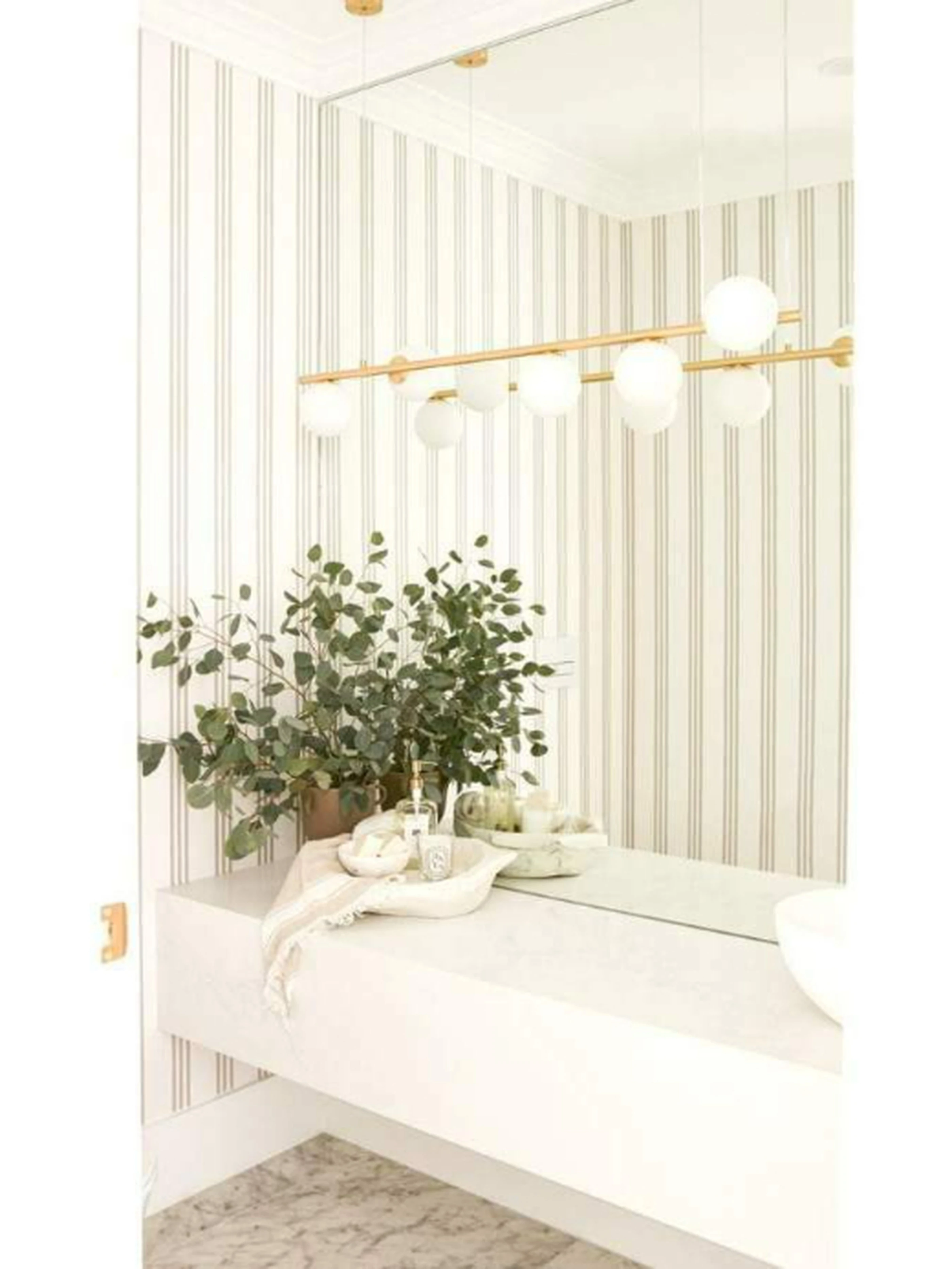 Modern bathroom with striped wallpaper, gold light fixture with white globes, floating vanity, and potted eucalyptus plant.