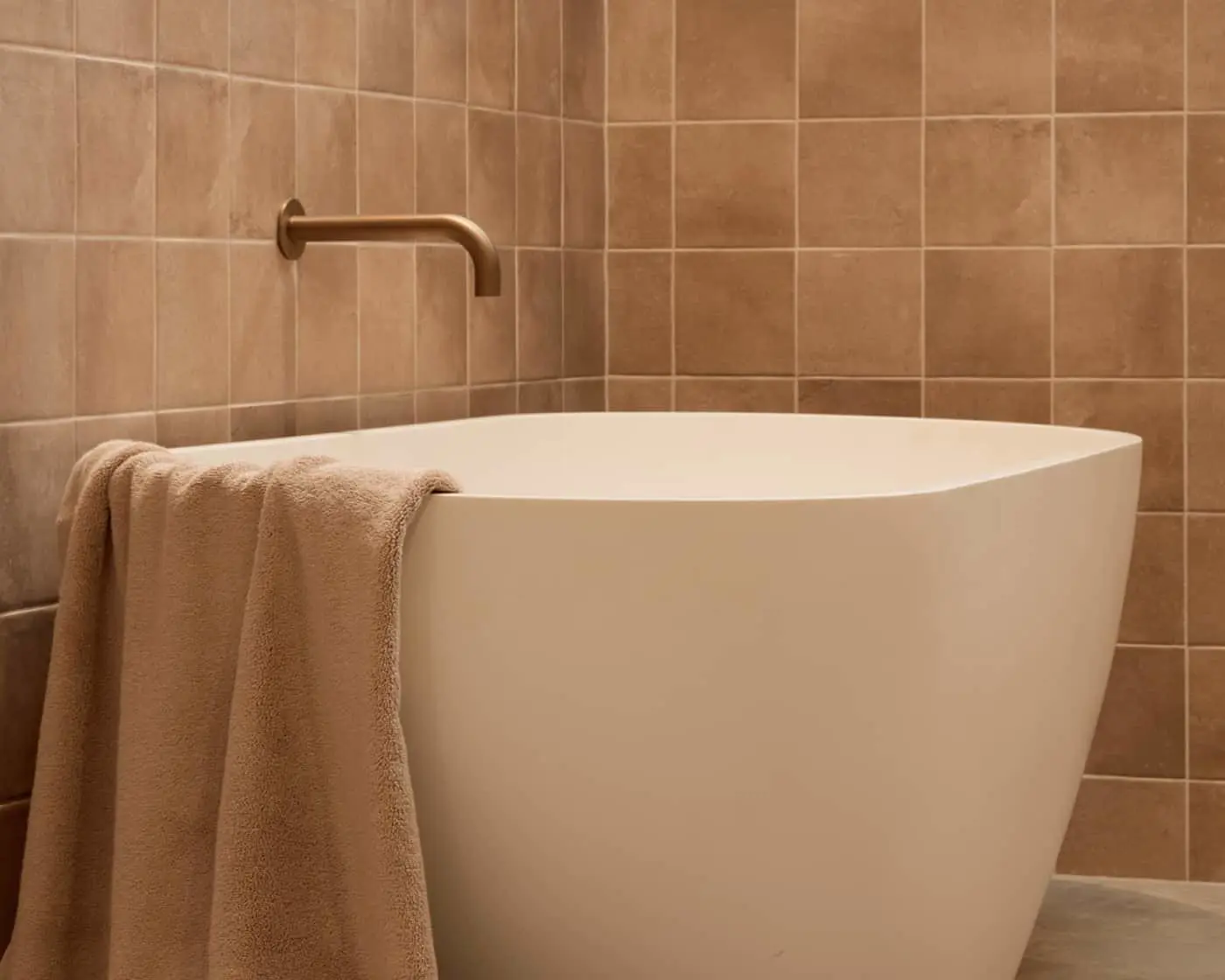 Modern white freestanding bathtub with beige towel and bronze wall-mounted faucet against brown tiled bathroom wall.