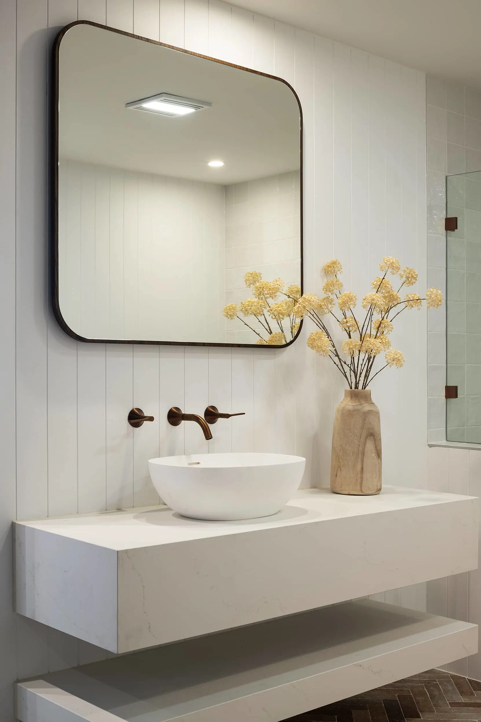 Modern bathroom with white vessel sink, bronze wall-mounted faucet, square mirror, and wooden vase with yellow flowers.