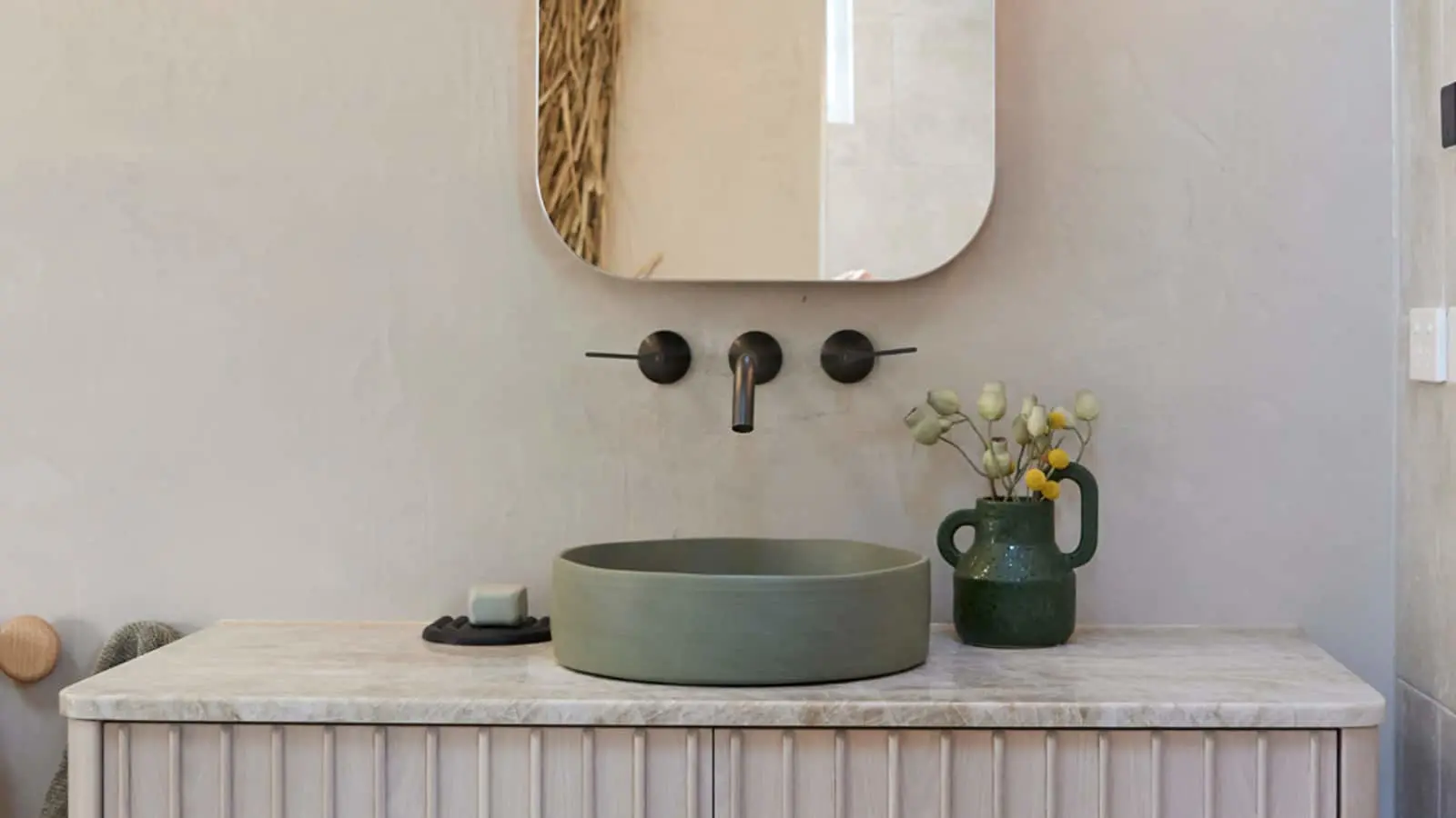Modern bathroom vanity with green vessel sink, wall-mounted black faucet, rounded mirror, and small green vase with flowers.
