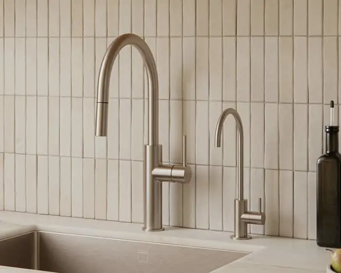 Two brushed nickel kitchen faucets on a white countertop with a vertical white tile backsplash and a dark glass bottle nearby.