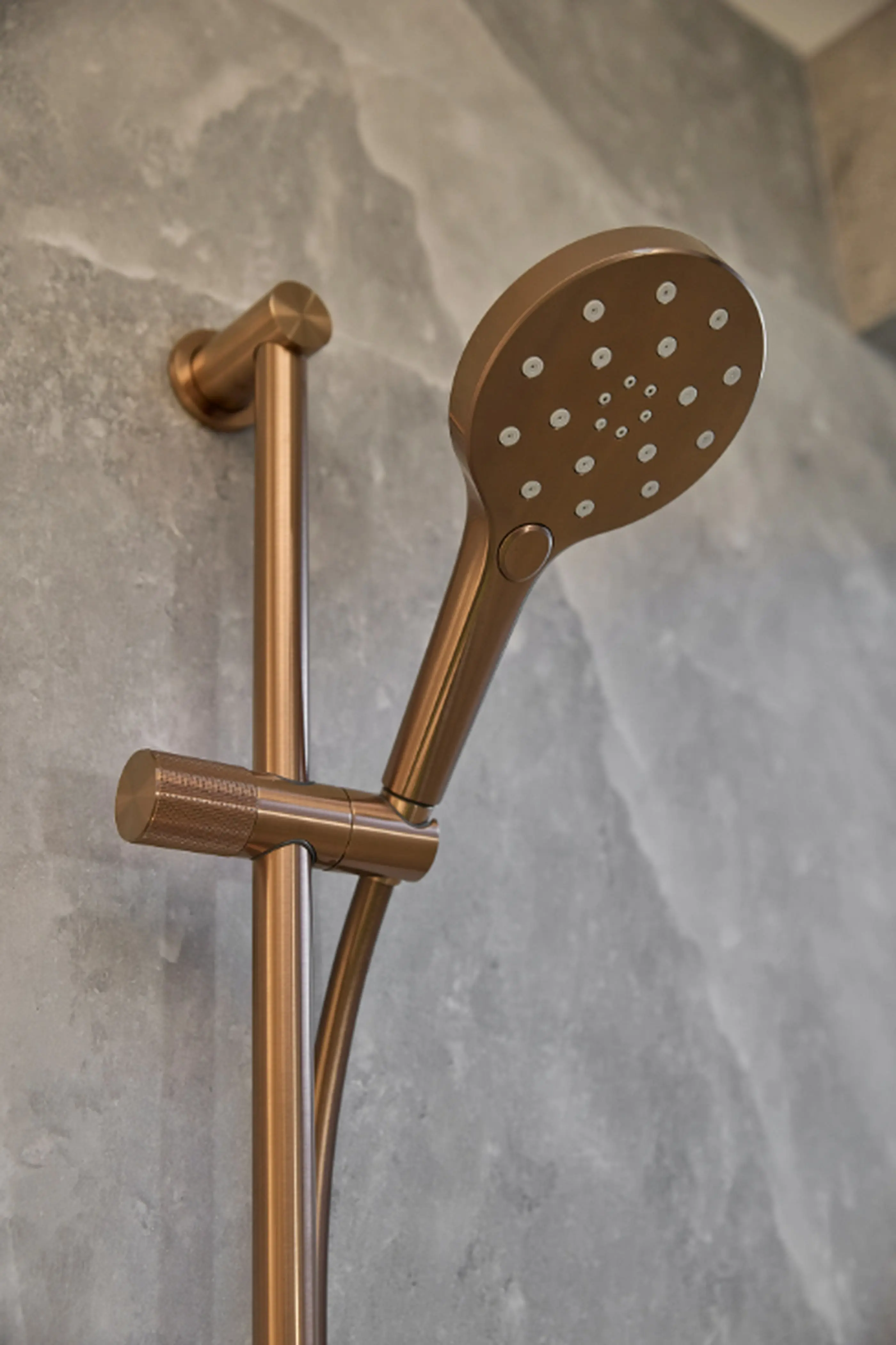 Bronze handheld shower head with multiple spray nozzles mounted on an adjustable rail against a gray stone wall.