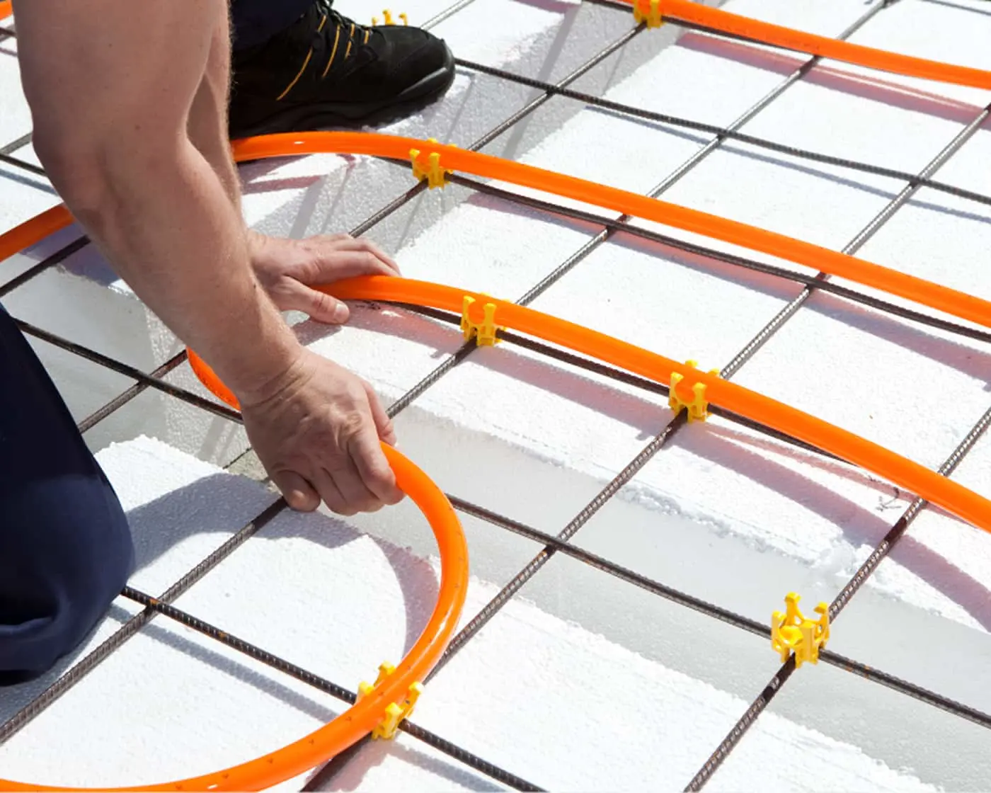 Worker installing orange radiant floor heating tubes on wire mesh grid with yellow clips.