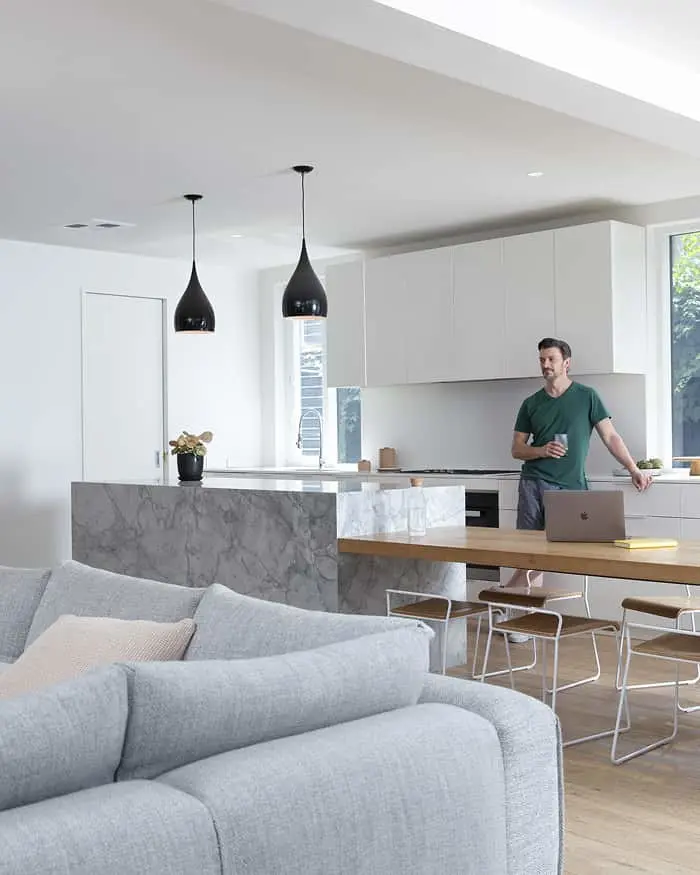 Modern open concept kitchen with marble island, black pendant lights, gray sofa, and person standing by laptop.