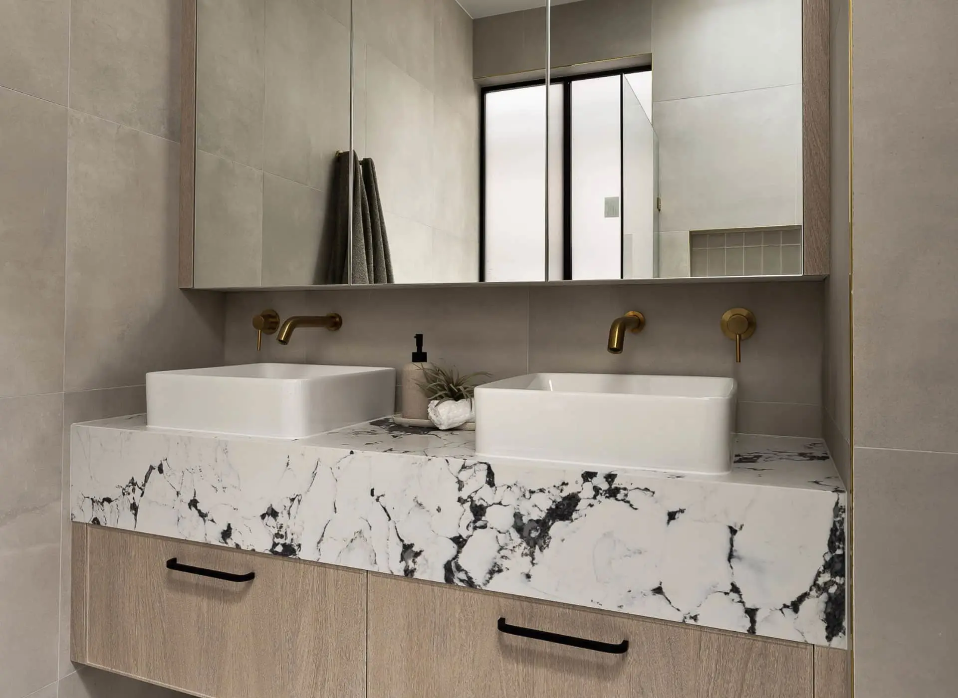 Modern bathroom with dual white vessel sinks on marble countertop, brass fixtures, and large mirror cabinet.