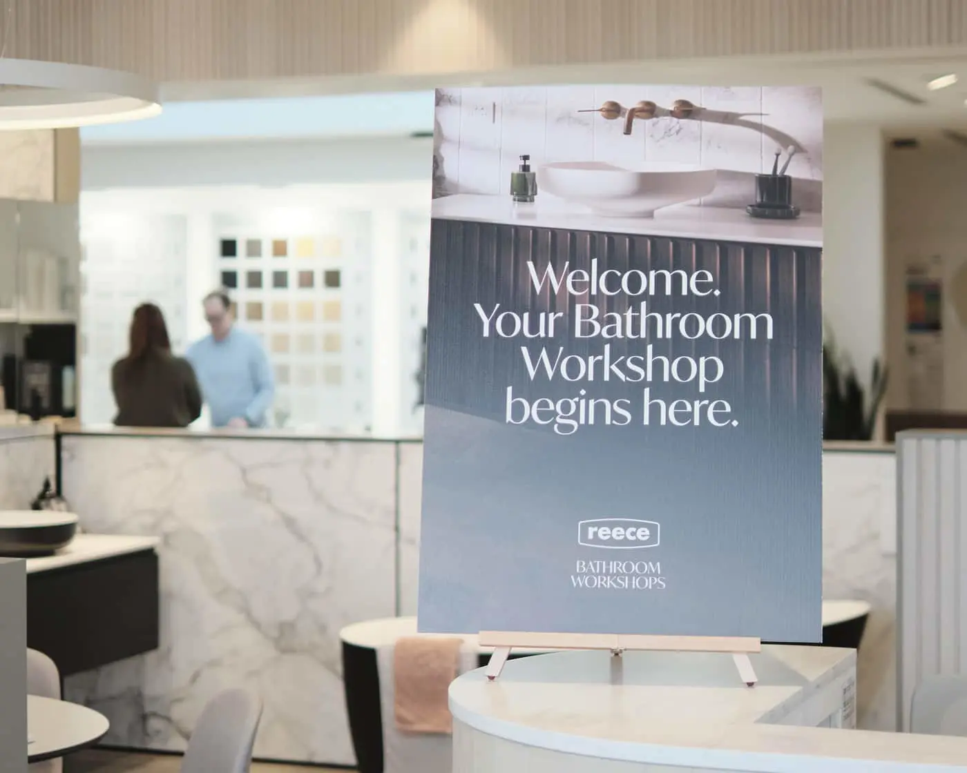 A Reece Bathroom Workshops welcome sign on an easel inside a showroom, with two people visible in the background.
