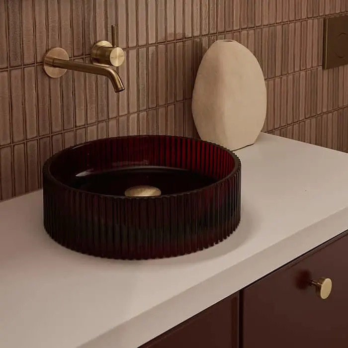 Modern bathroom with deep red ribbed glass vessel sink, brass wall-mounted faucet, and beige tiled wall.