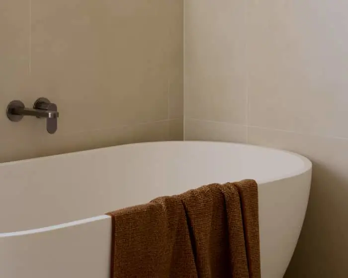 White freestanding bathtub with brown towel draped over edge and dark wall-mounted faucet in tiled bathroom