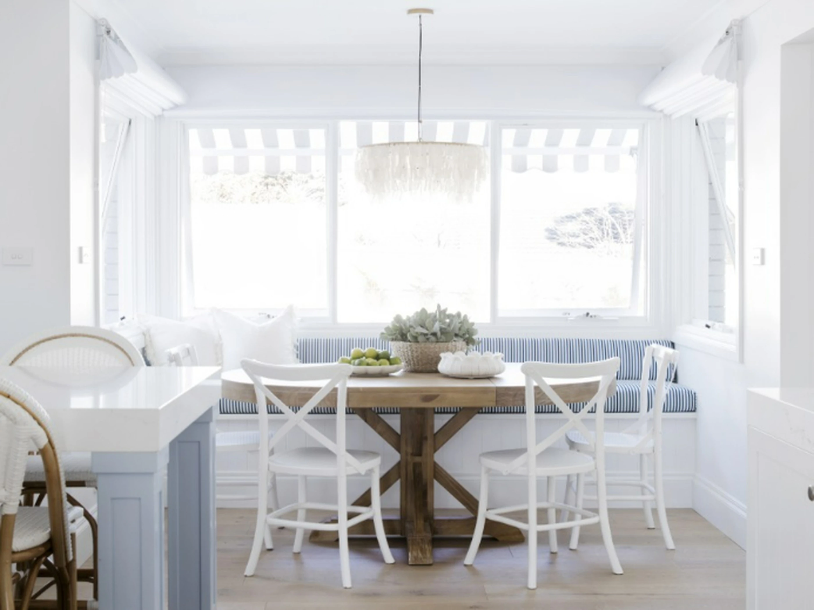 Bright coastal dining nook with wooden table, white chairs, blue striped bench seating, and shell chandelier by large windows.