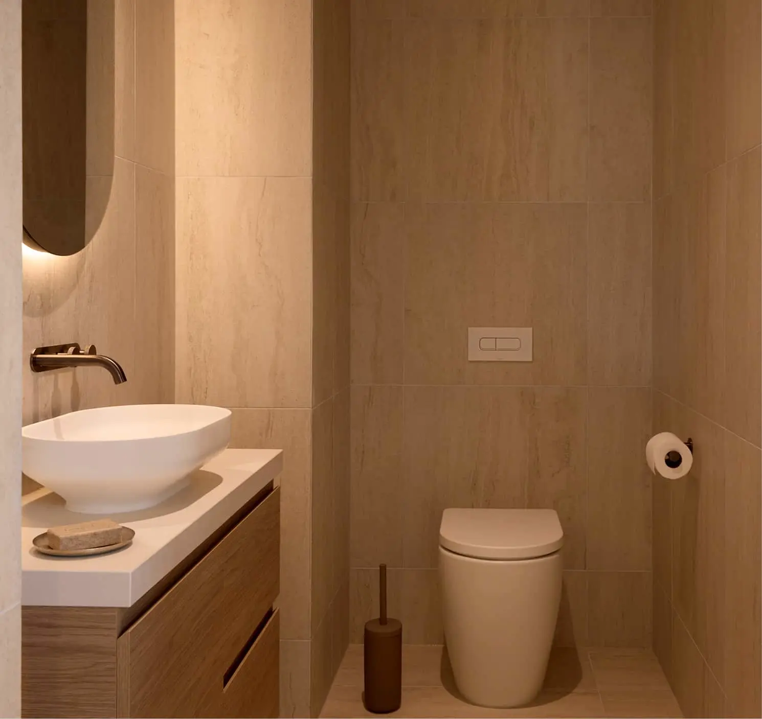 Small bathroom with beige stone tiles, a white vessel sink on a wood vanity, wall-mounted faucet, round mirror, and a toilet.