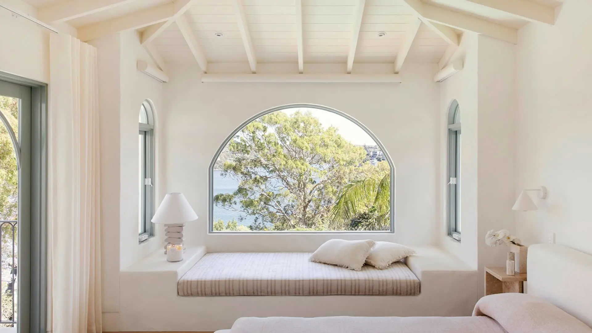 White bedroom with built-in window seat beneath arched window overlooking trees and water, featuring beamed ceiling.