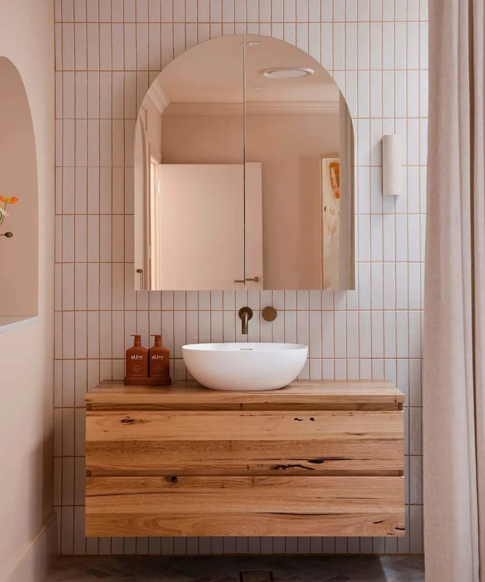 Modern bathroom with white vertical tiles, arched mirror, wooden vanity, white vessel sink, and brass fixtures.