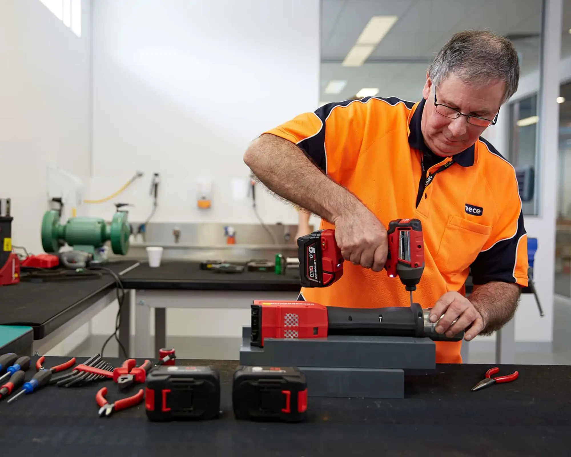 Worker in orange safety shirt using a red power drill on equipment in a workshop with tools on the workbench.