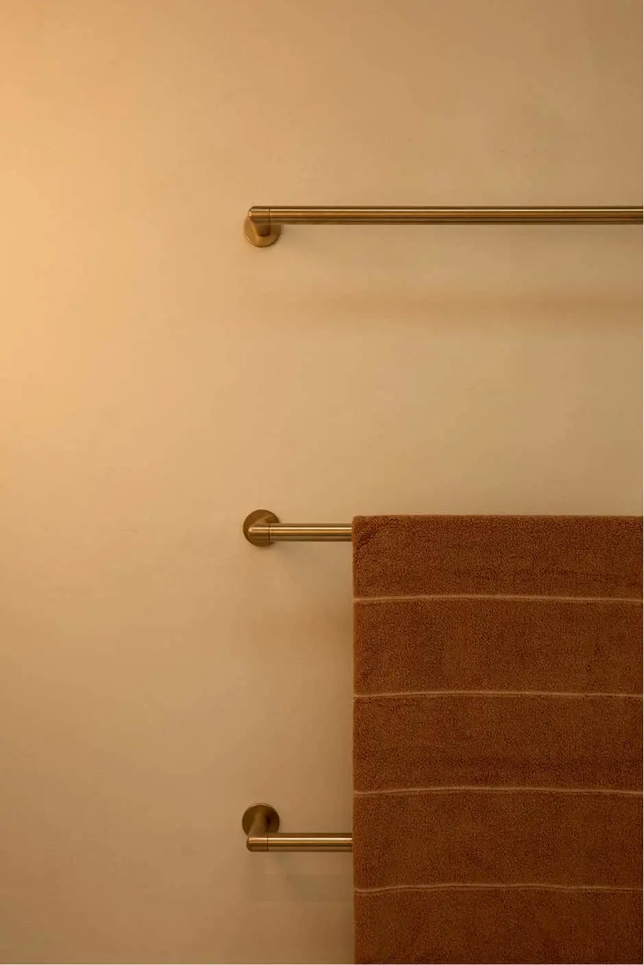 Three brass towel bars mounted on a beige wall with a brown towel hanging on the lower bar.