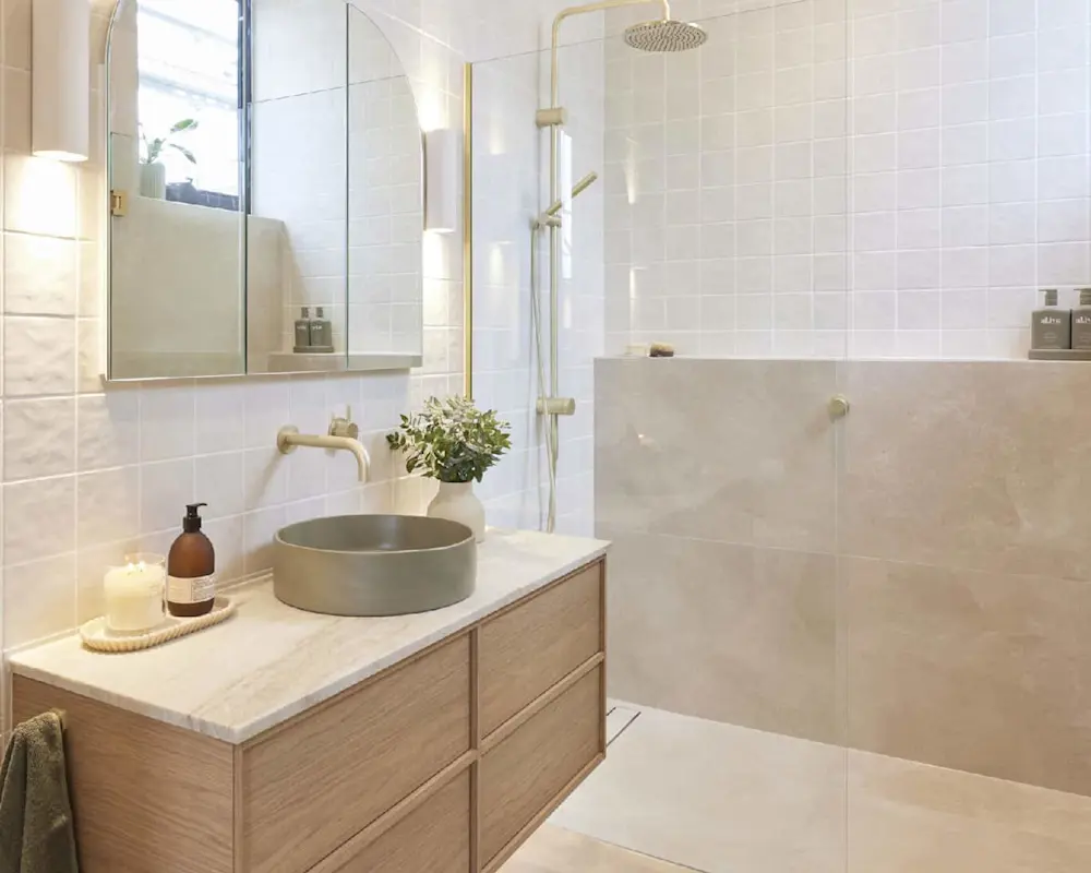 Modern bathroom with white tiles, wooden vanity, gray vessel sink, gold fixtures, and glass shower with rainfall head.
