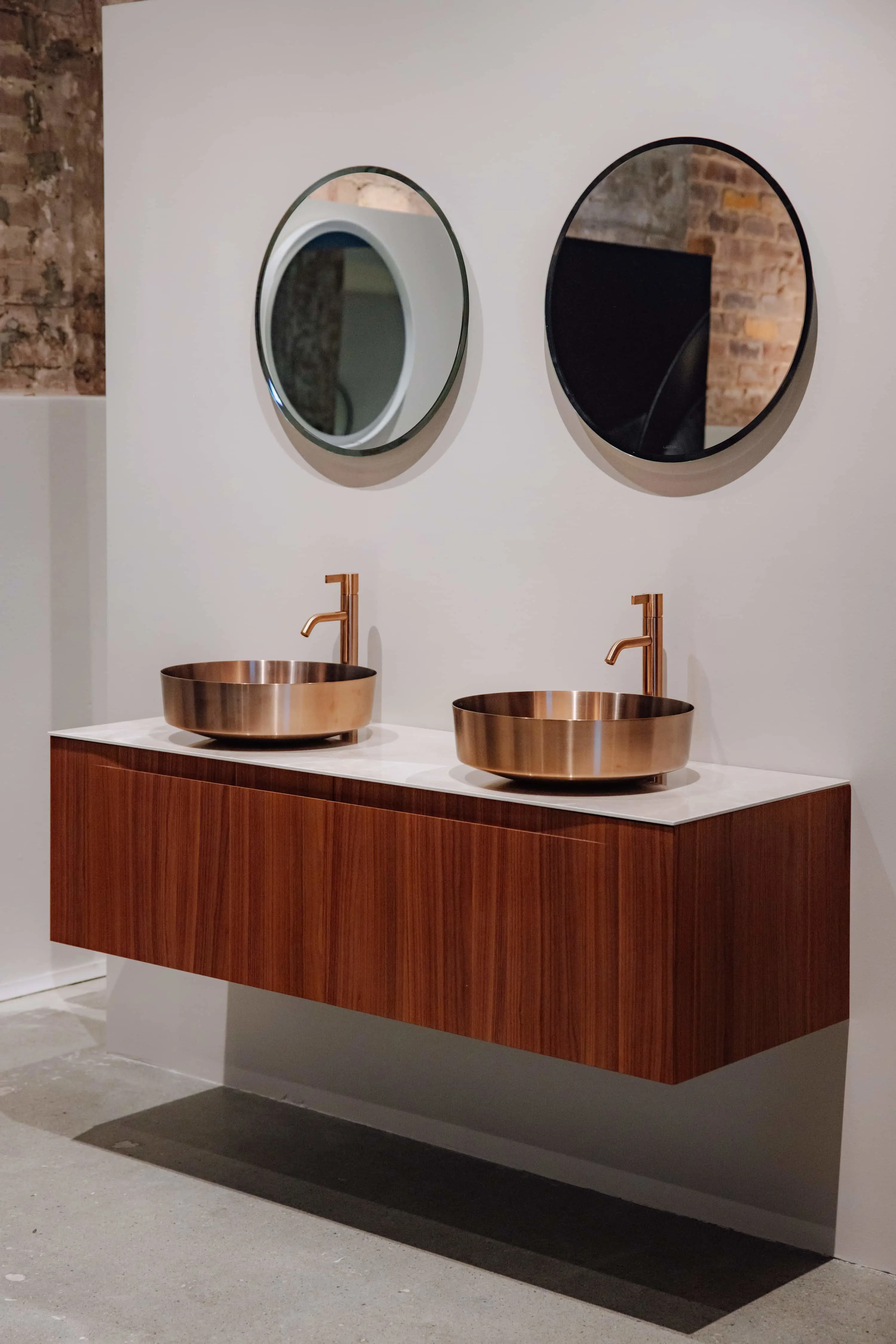 Modern bathroom with floating wooden vanity, copper vessel sinks, brass faucets, and two round mirrors against exposed brick wall.