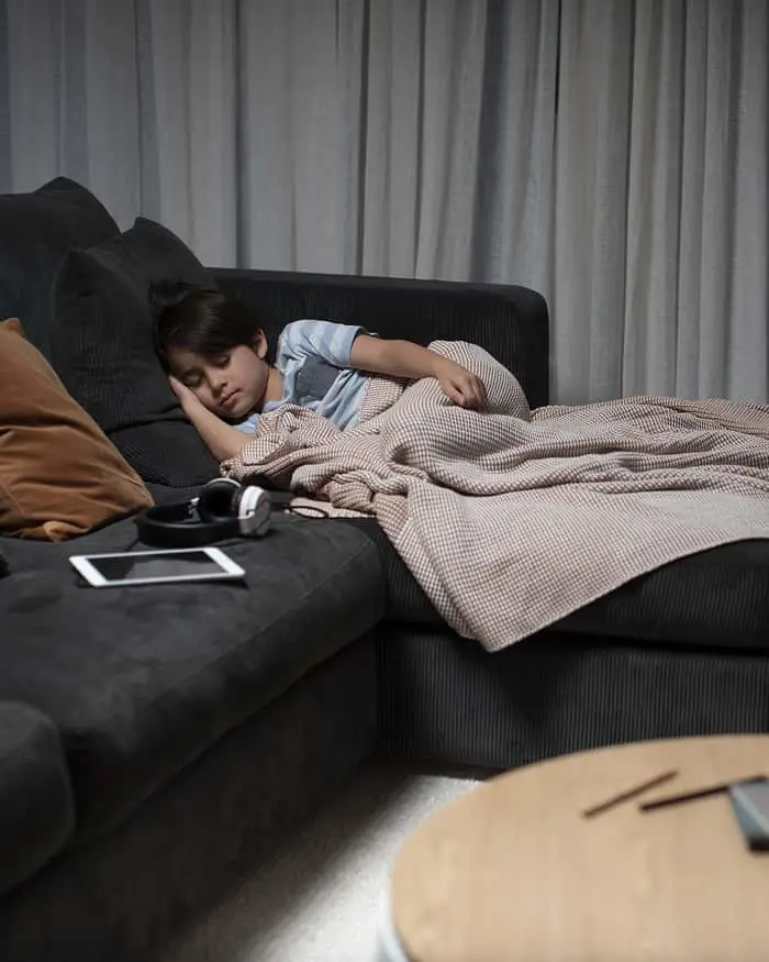 Child sleeping on dark gray couch covered with beige blanket, smartphone and headphones on nearby ottoman.