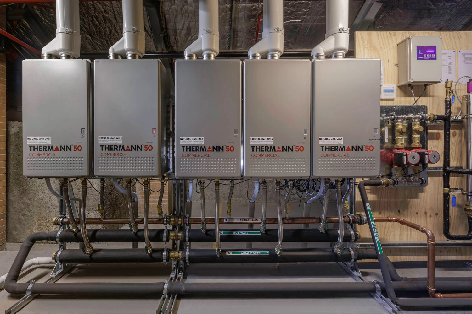 Five Thermann 50 Commercial natural gas water heaters installed in a row with connected piping system in a utility room.