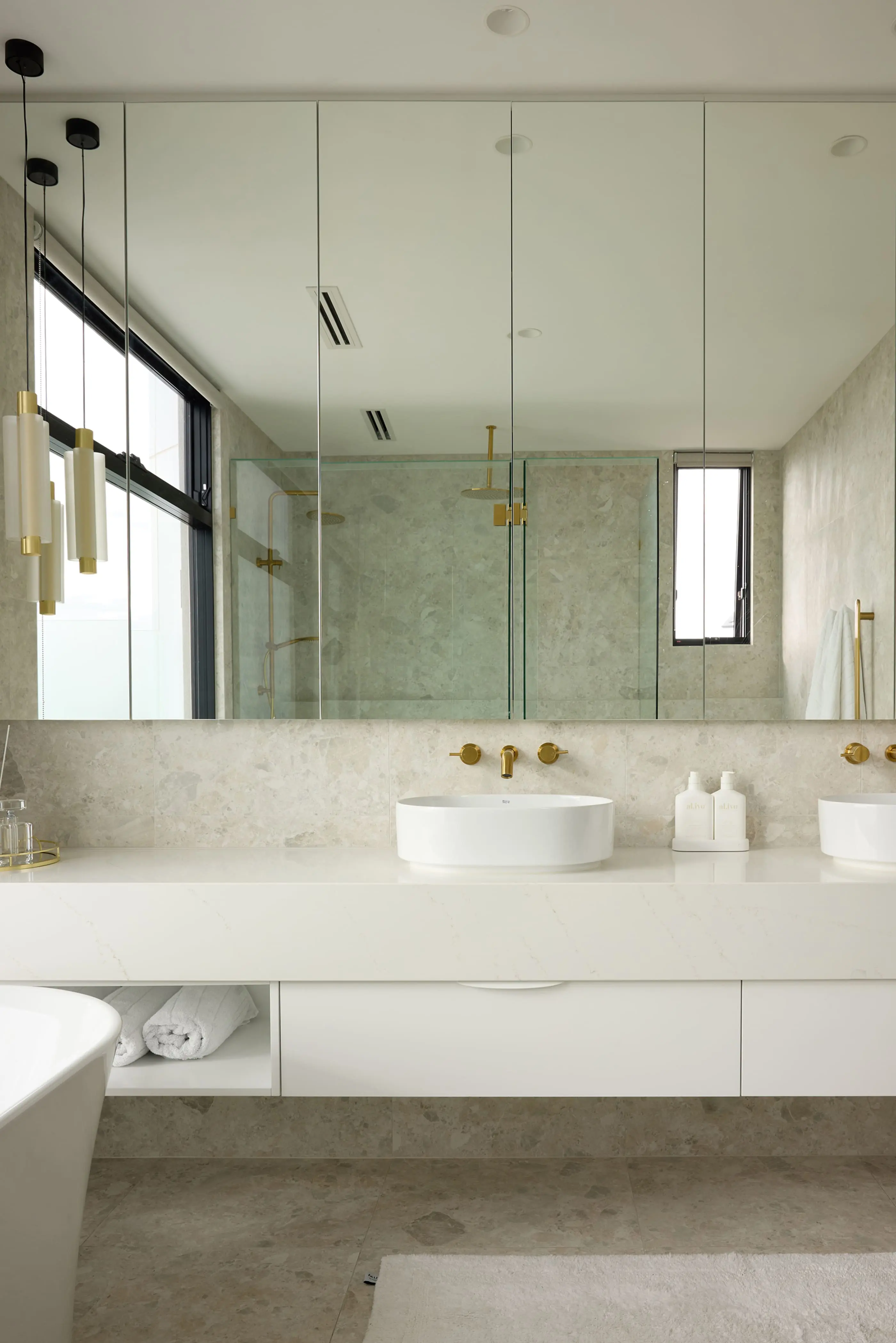Modern bathroom with white vessel sink, marble countertop, brass fixtures, and large mirror reflecting glass shower enclosure.