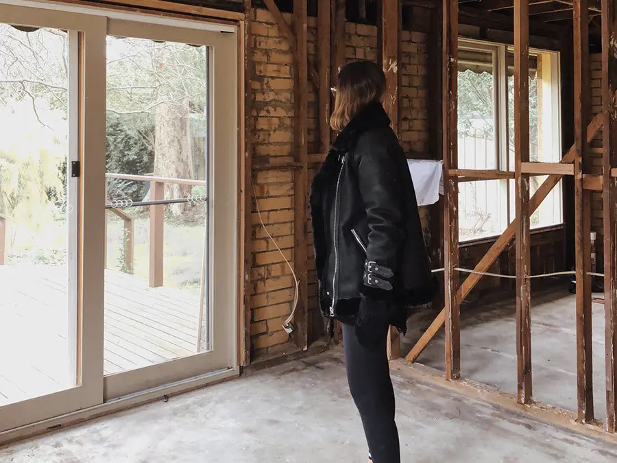 Person in black jacket looking out glass doors in home under renovation with exposed brick walls and wooden framing.