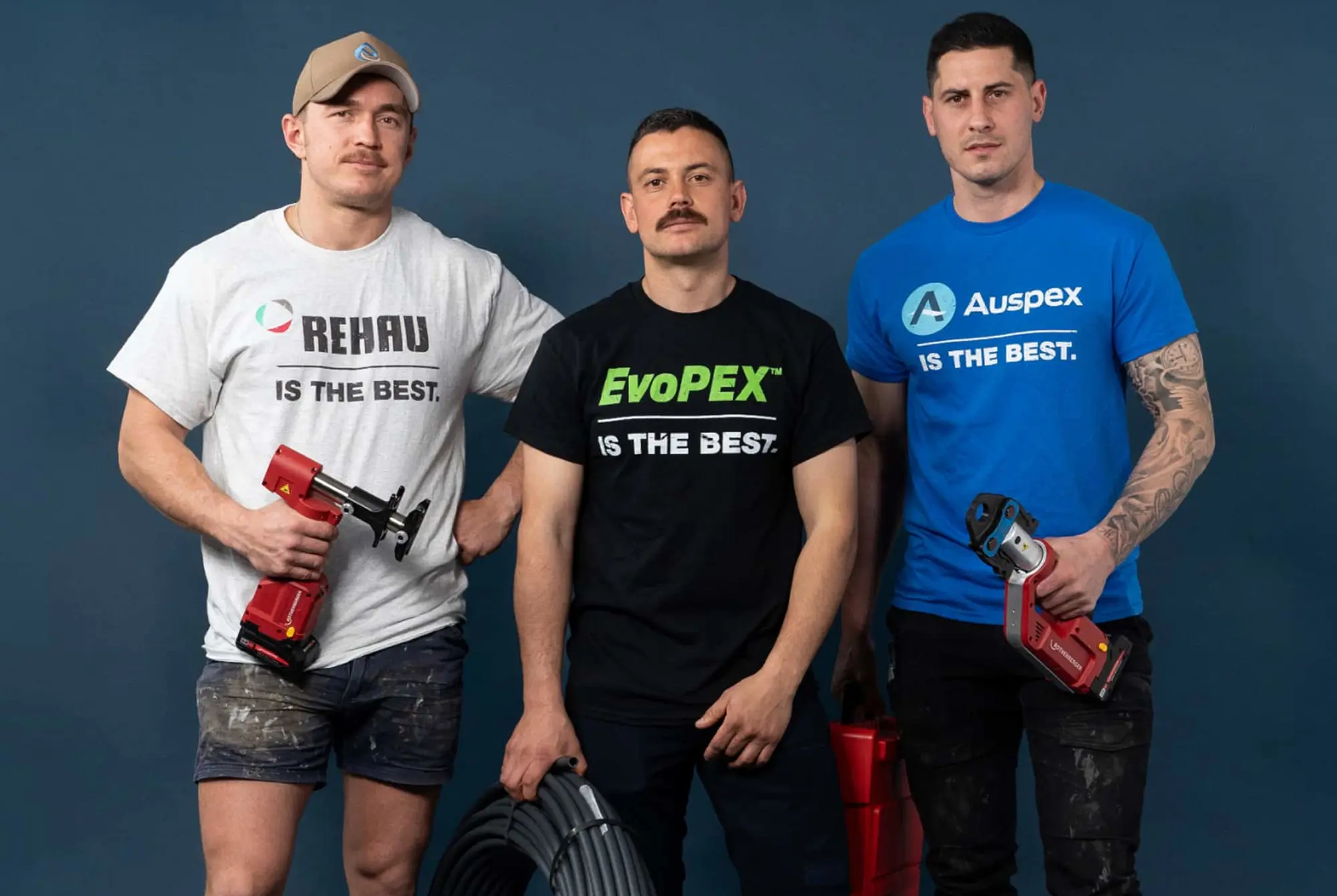 Three tradesmen standing against blue background wearing branded t-shirts and holding plumbing tools and pipe.