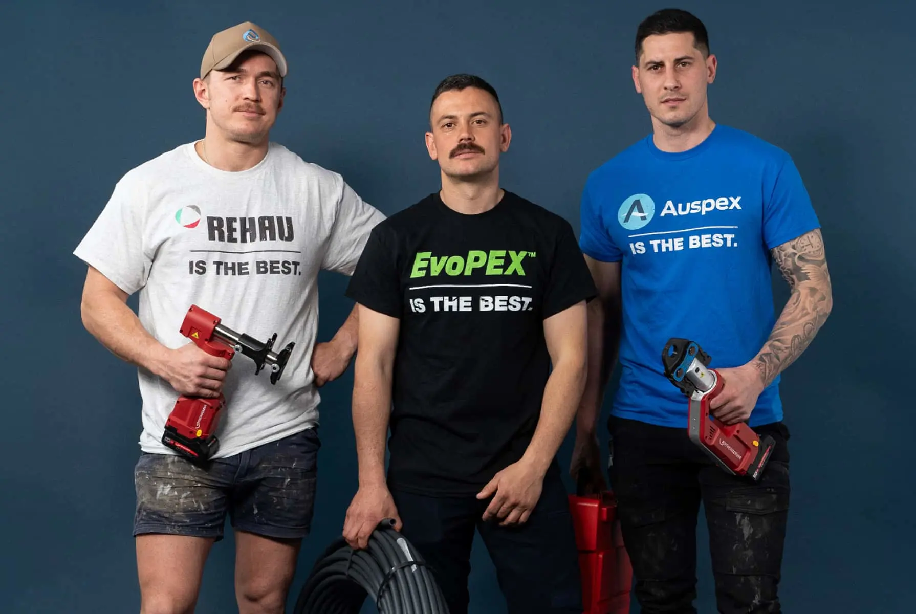 Three tradesmen standing against blue background wearing branded t-shirts and holding plumbing tools and pipe.