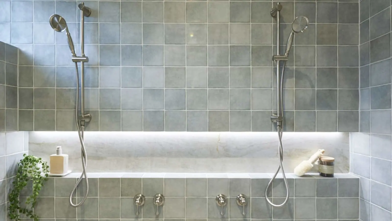 Modern walk-in shower with blue-gray tiles, dual showerheads, built-in shelf with toiletries, and small plant.