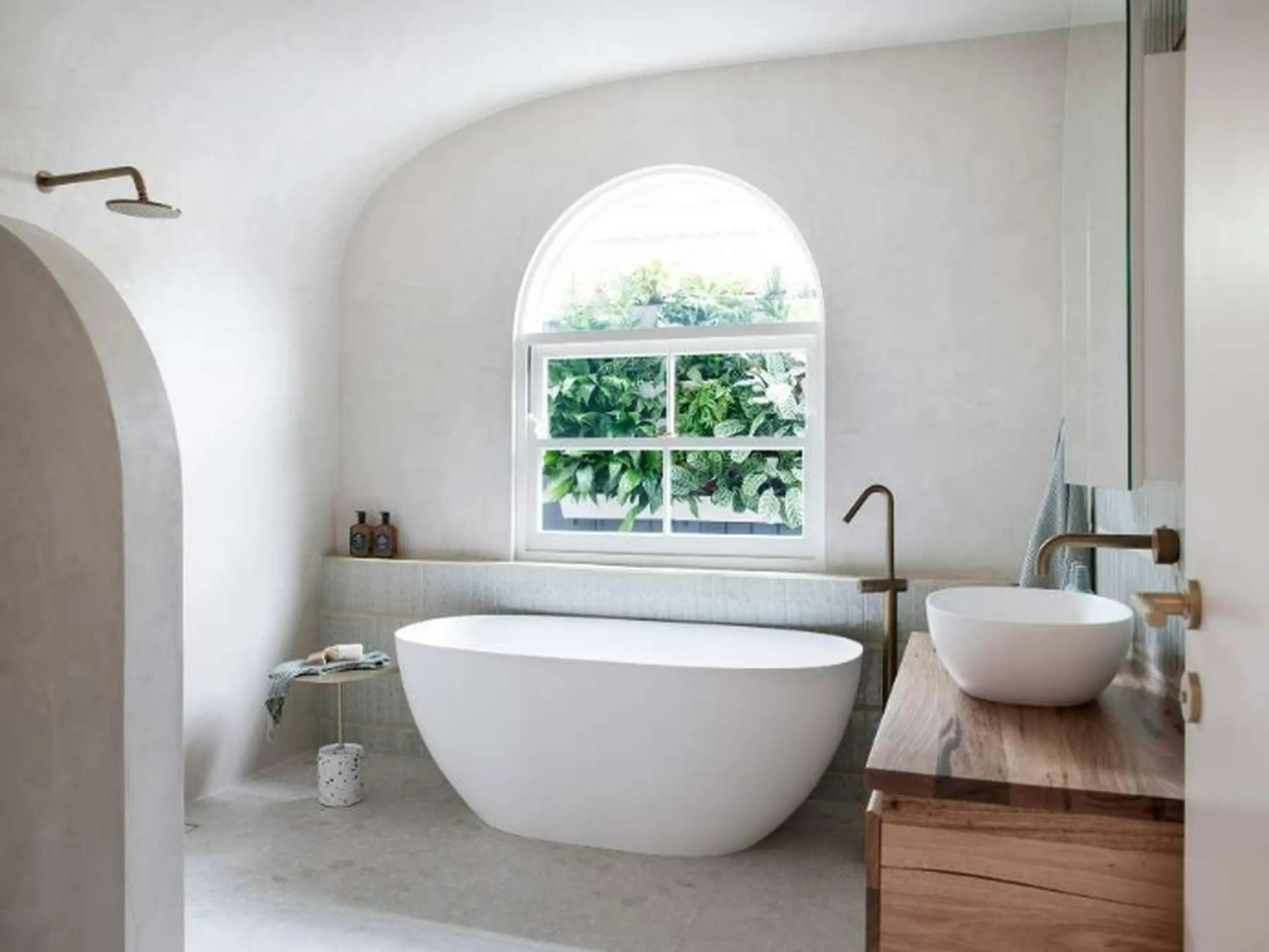 Modern white bathroom with freestanding tub, wooden vanity, and arched window overlooking greenery.