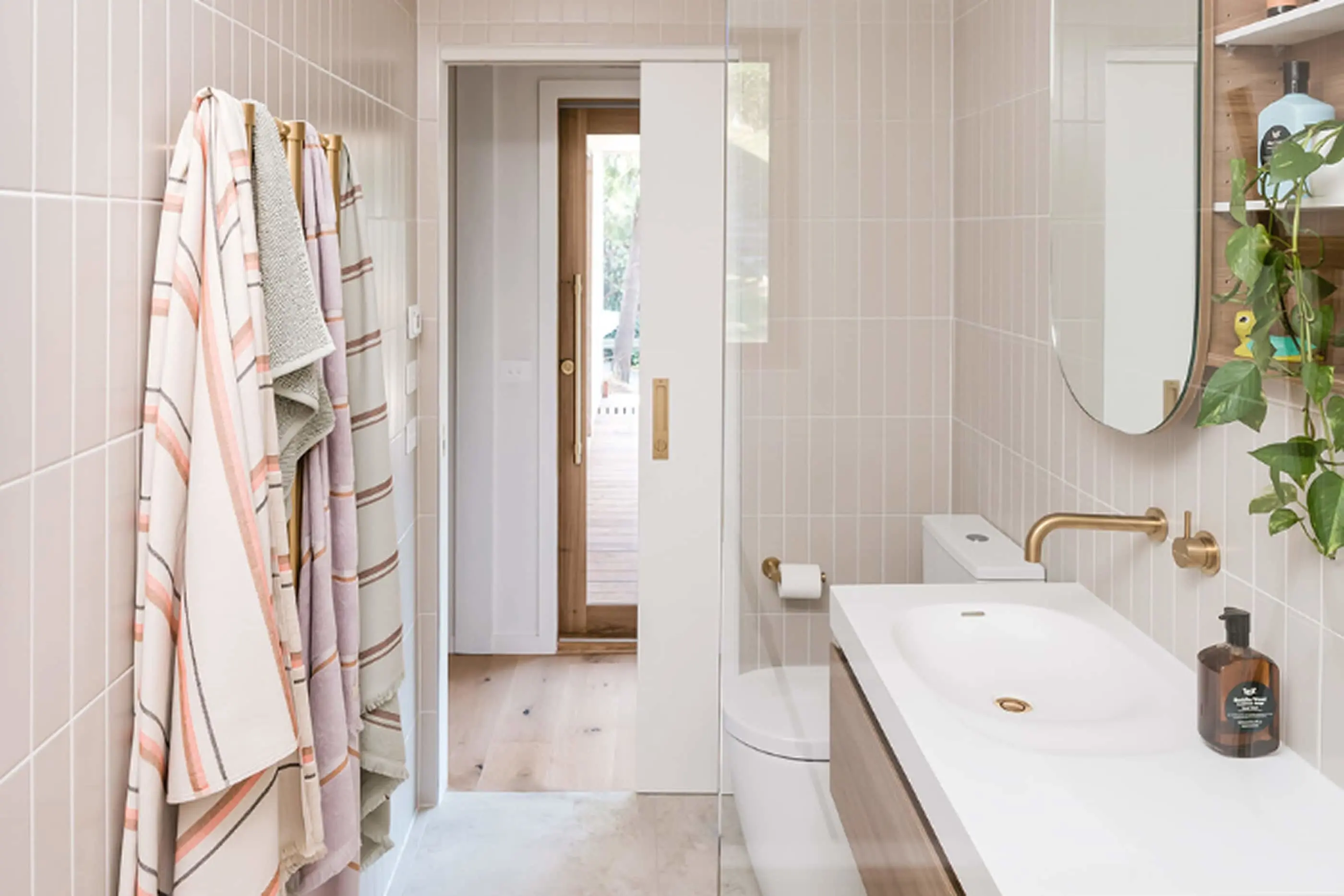Modern bathroom with white tile walls, hanging towels, white sink with brass fixtures, round mirror, and potted plant.