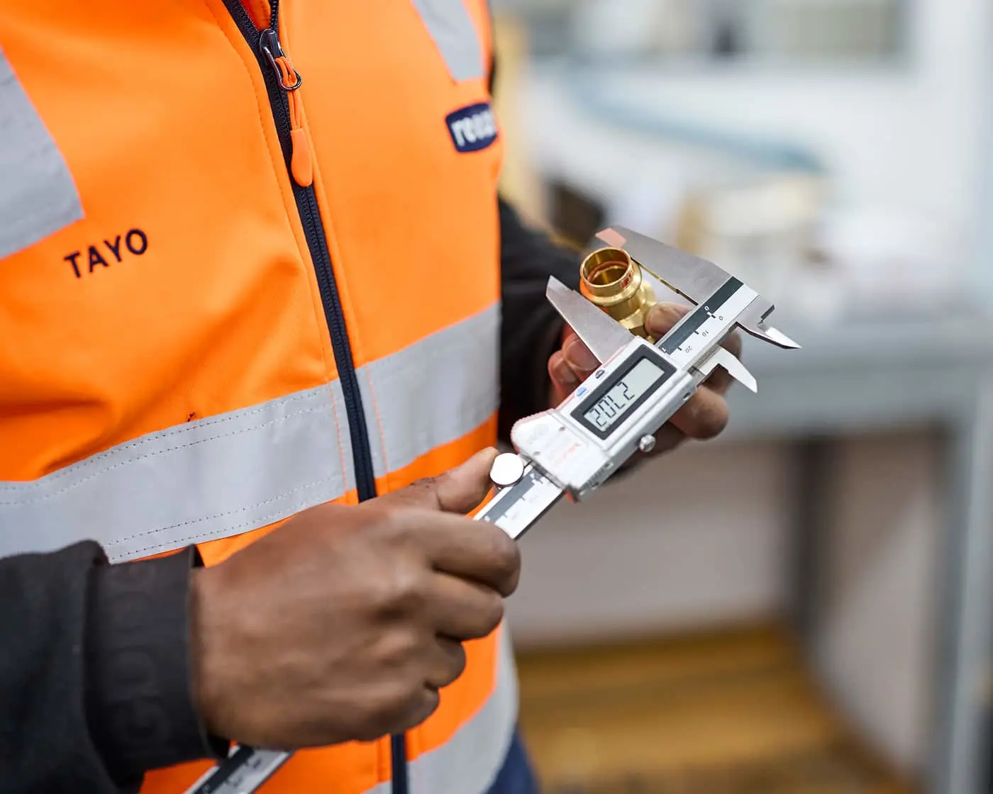 Worker in orange TAYO safety vest measuring a brass fitting with digital calipers at a construction site.