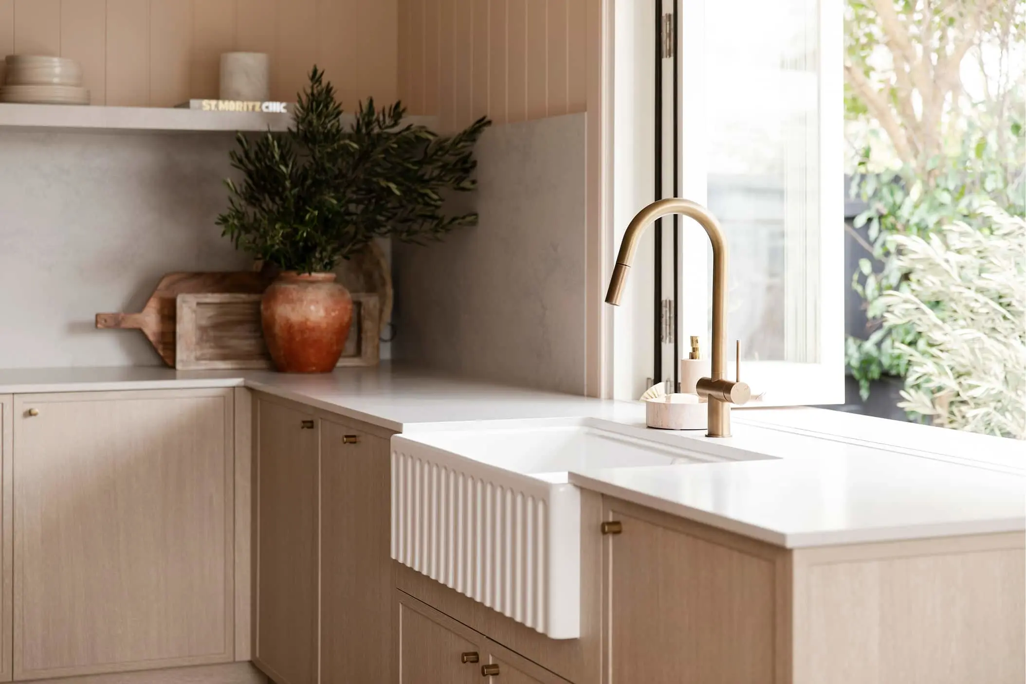 Modern kitchen with farmhouse sink, brass faucet, light wood cabinets, and decorative plant in terracotta pot by window.