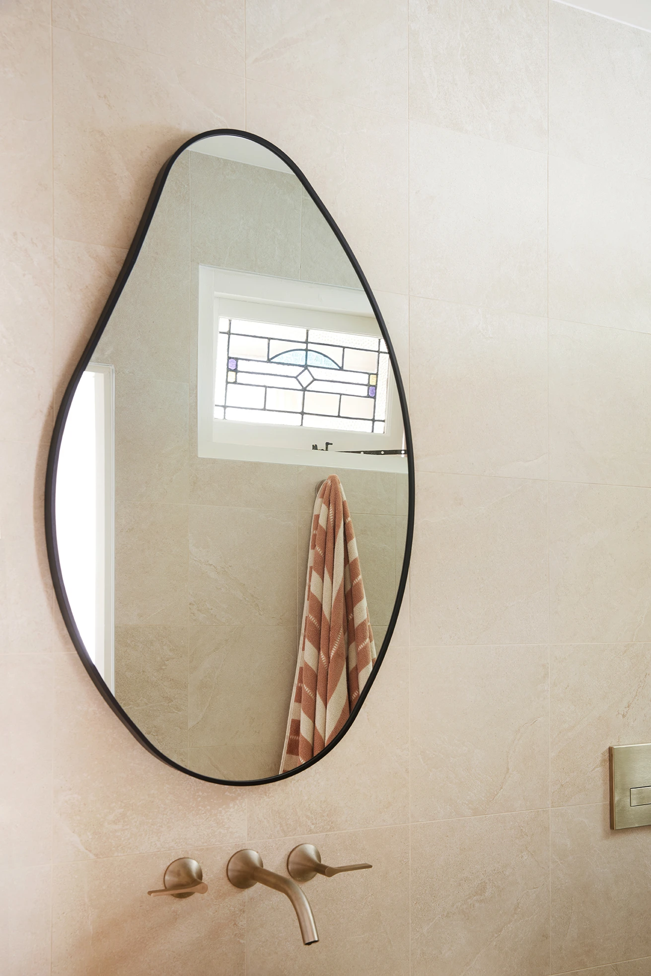 An organic-shaped mirror with a black frame on a beige tiled bathroom wall above brushed gold wall-mounted taps.