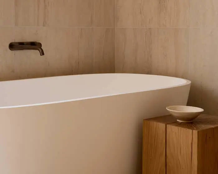 A white freestanding bathtub against beige stone tiles, with a bronze wall-mounted faucet and a small wooden side table holding a bowl.