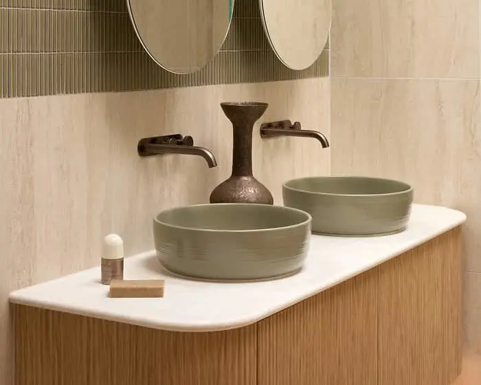 Two green ceramic vessel sinks on a white countertop with bronze wall-mounted faucets and a decorative vase between them.