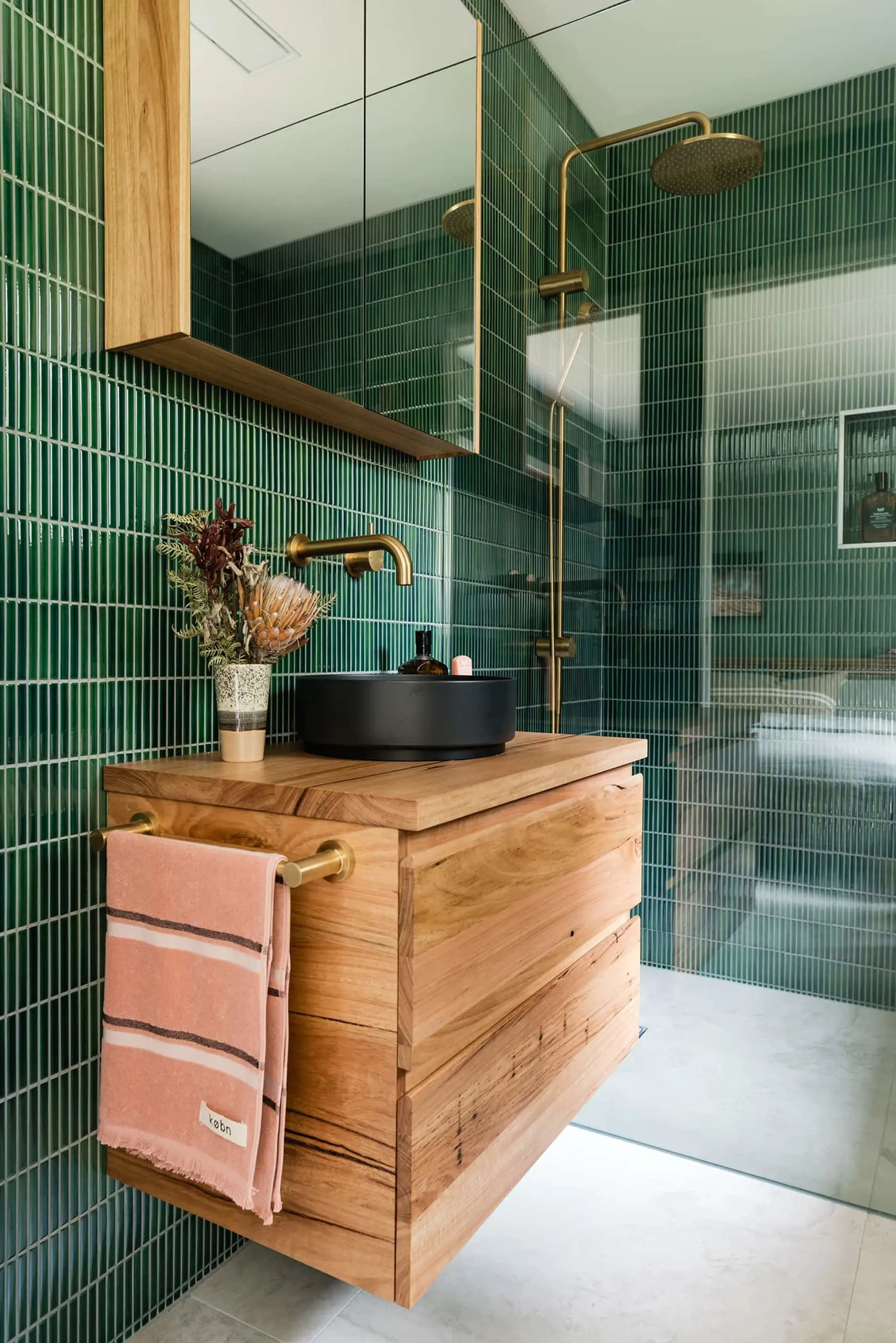 Modern bathroom with green tile walls, wooden vanity with black basin, brass fixtures, and glass shower enclosure.
