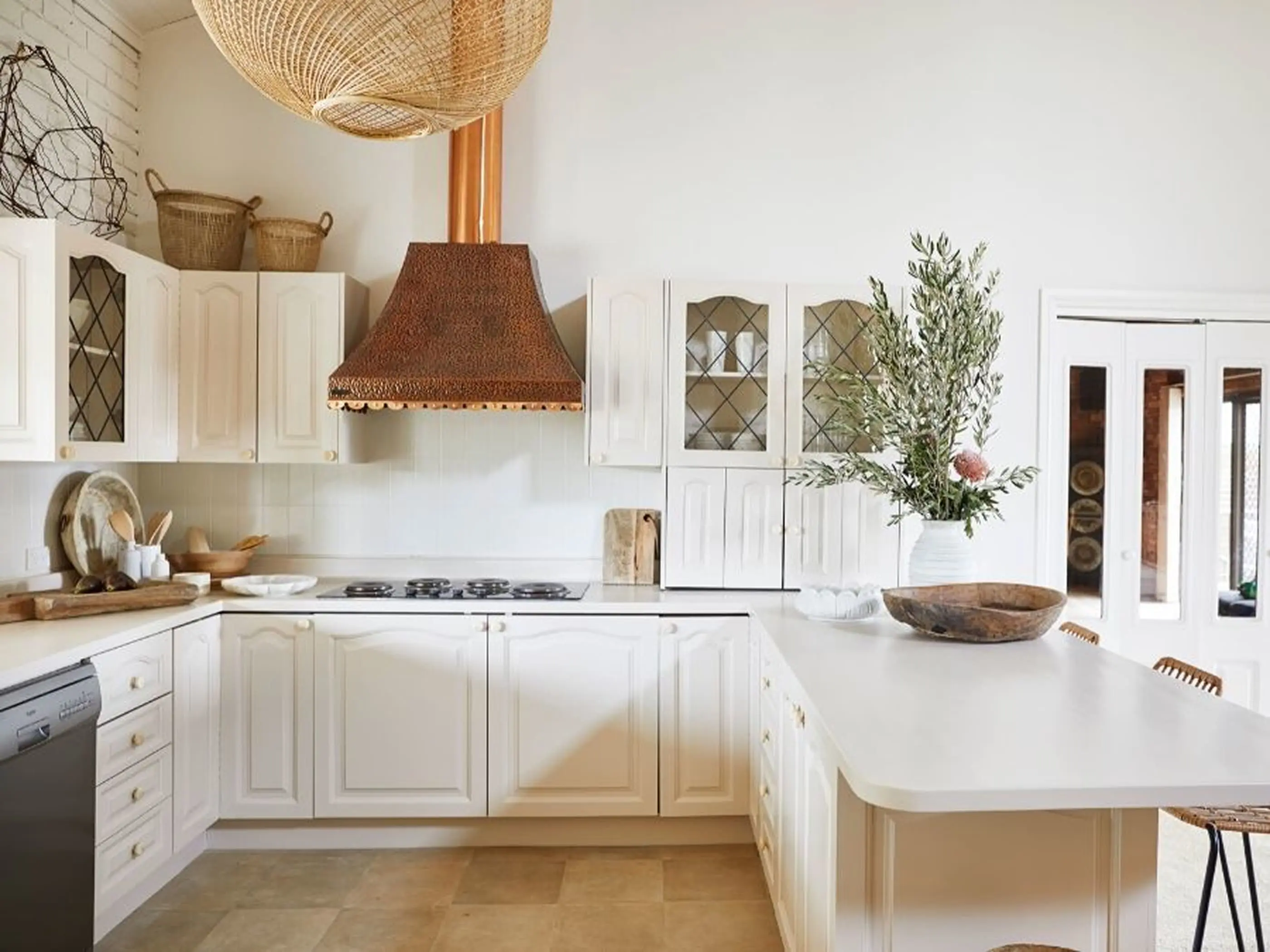 White kitchen with copper range hood, woven pendant light, white cabinets, and greenery in a vase on the counter.
