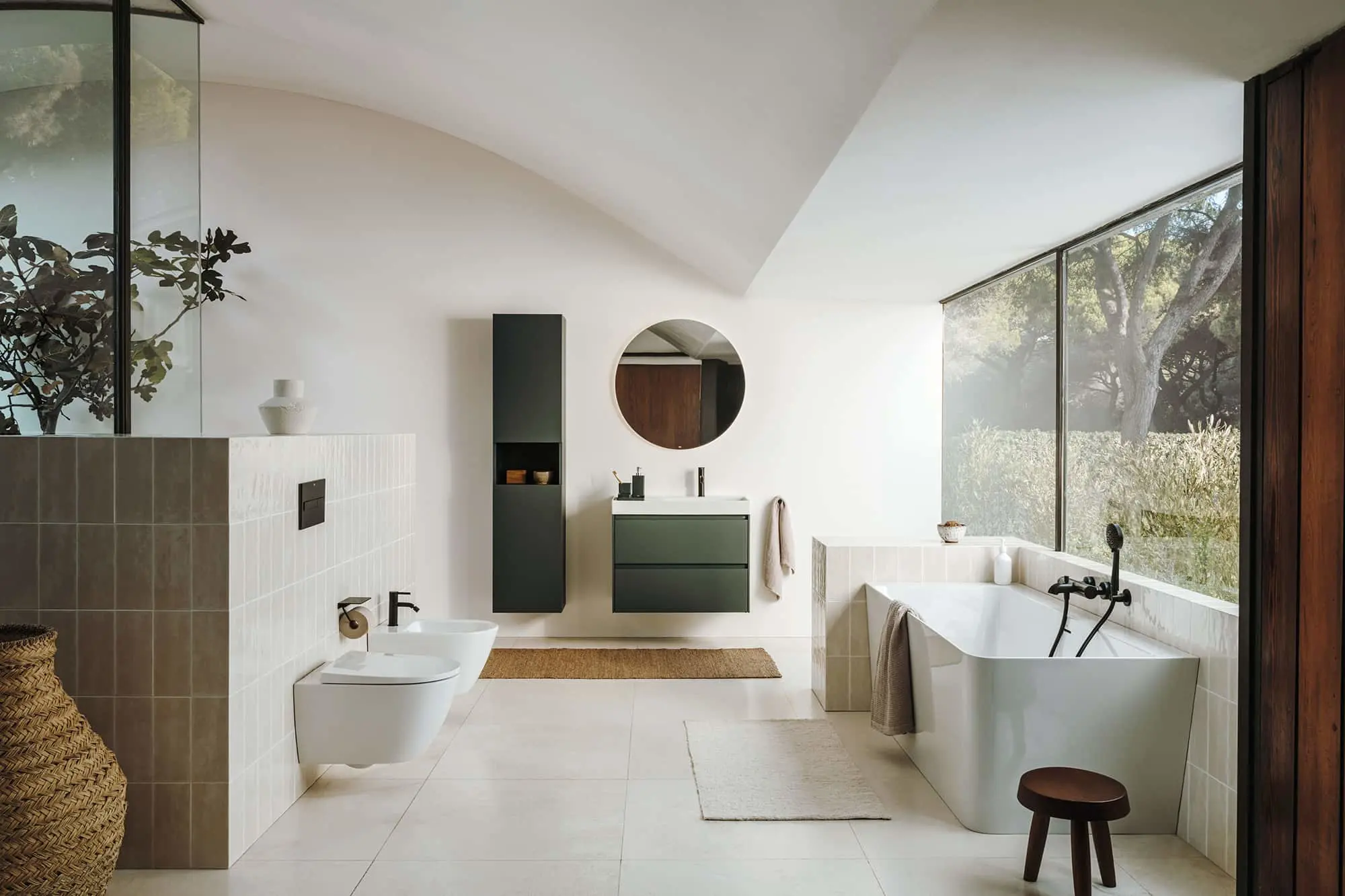 Modern bathroom with white freestanding tub, green vanity, round mirror, and large windows overlooking garden.