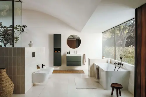 Modern bathroom with white freestanding tub, green vanity, round mirror, and large windows overlooking garden.