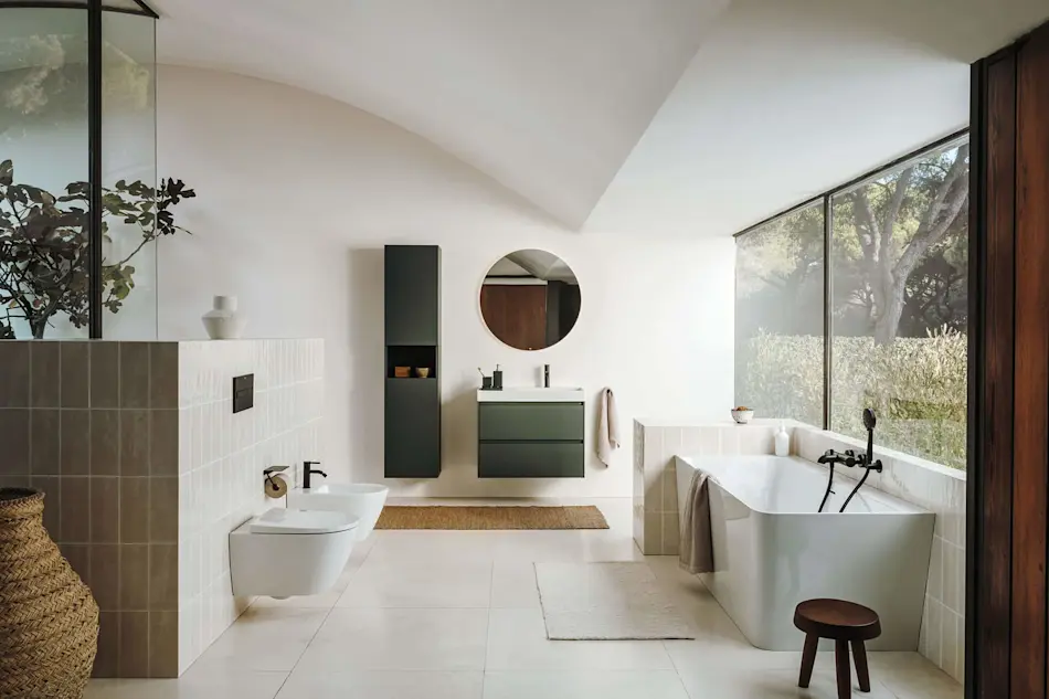 Modern bathroom with white freestanding tub, green vanity, round mirror, and large windows overlooking garden.