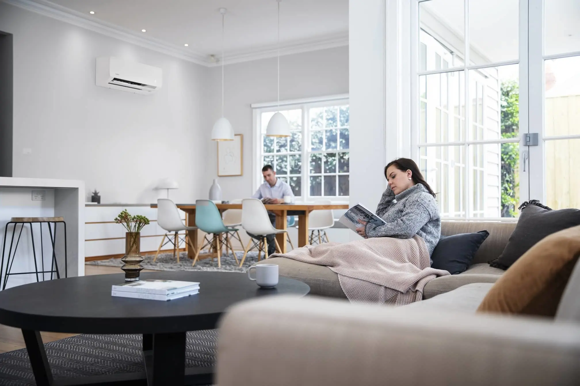 Person reading on couch in bright modern living room with dining area and another person working at table in background.